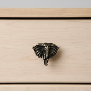 May include: A bronze-colored elephant-shaped drawer pull is mounted on a light-colored wooden drawer. The drawer pull is detailed with the elephant's head, trunk, and ears. The wood grain is visible on the drawer.