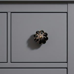 May include: Close-up of a gray cabinet drawer with a black and gold flower-shaped knob. The knob has a glossy finish and a detailed design. The cabinet is painted in a solid gray color.
