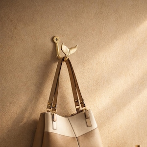 May include: A gold-colored wall hook with a whale tail design, holding a beige and brown handbag. The hook is mounted on a textured, light brown wall. The handbag has two long straps and gold-colored hardware.