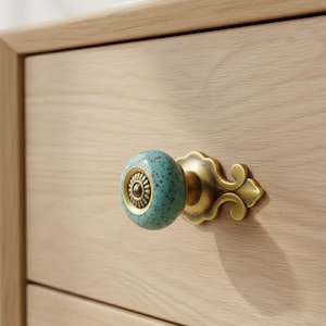 May include: Close-up of a drawer knob with a teal speckled ceramic handle and an ornate brass base. The knob is mounted on a light-colored wooden drawer, showcasing a blend of textures and colors.