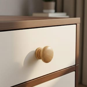 May include: Close-up of a white drawer with a light brown wooden knob. The drawer is part of a larger piece of furniture with a dark brown trim and top. The knob is oval-shaped and casts a shadow on the drawer.