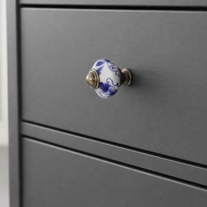 May include: A decorative cabinet knob with a white and blue floral design. The knob has a brass base and is mounted on a dark gray cabinet. The knob is oval-shaped and features a detailed pattern.