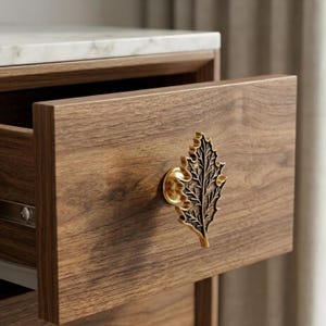 May include: A wooden drawer with a decorative leaf-shaped brass handle. The drawer is open, revealing the interior. The wood grain is visible, and the countertop above is white marble. The handle is detailed with intricate leaf veins.