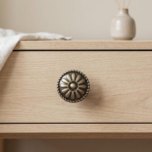 May include: A bronze-colored drawer pull with a floral design and a rope-like border. The hardware is mounted on a light-colored wooden drawer. A small, off-white vase and a folded white cloth are visible in the background.
