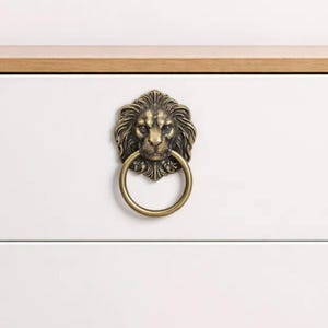 May include: Antique brass lion head drawer pull with a ring handle. The lion's face is detailed with a textured mane and a stern expression. The pull is mounted on a white drawer, with a wooden top in the background.