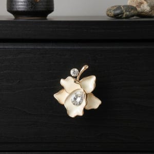 May include: A decorative cabinet knob in the shape of a flower. The knob features a cream-colored enamel finish with gold accents and is adorned with two clear, sparkling crystals. The flower knob is mounted on a black surface.