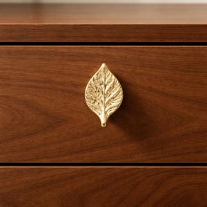 May include: A gold-colored leaf-shaped drawer pull mounted on a dark brown wooden drawer. The leaf has detailed veining and a textured surface. The drawer is part of a larger piece of furniture.