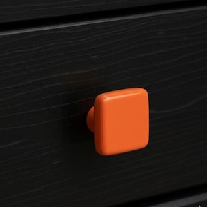 May include: Close-up of a black wooden drawer with an orange square knob. The knob is centered on the drawer and has a glossy finish. The wood grain is visible in the black finish.