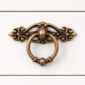May include: Close-up of an antique brass drawer pull with a ring handle. The decorative hardware features ornate scrollwork and a warm, aged finish. The pull is mounted on a white surface.
