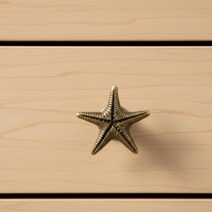 May include: A bronze-colored starfish cabinet knob is mounted on a light wood drawer. The five-pointed starfish has detailed ridges and a vintage finish. The drawer has a natural wood grain and horizontal black lines.