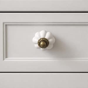 May include: White ceramic cabinet knob with a flower-like design and a brass-colored base. The knob is mounted on a white cabinet door. The hardware adds a decorative touch to furniture.