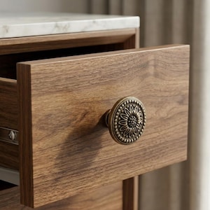 May include: A wooden drawer with a decorative brass sunflower knob. The drawer is open, revealing the interior. The cabinet has a marble top. The wood grain is visible on the drawer front.