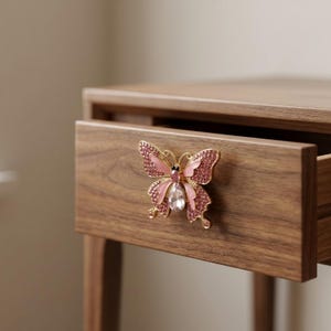 May include: A decorative butterfly drawer pull with pink and clear crystal accents and gold-colored metal detailing. The butterfly is mounted on a wooden drawer, which is partially open, revealing the interior.