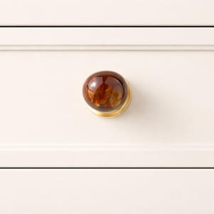 May include: A round cabinet knob with a brown, marbled appearance and a brass base. The knob is attached to a white cabinet door. This decorative hardware adds a touch of elegance to furniture.