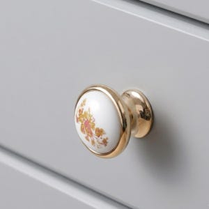 May include: A close-up of a white and gold cabinet knob with a floral design. The knob has a gold base and a white, rounded top with a gold rim. The floral pattern features pink and yellow flowers.