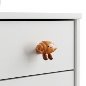 May include: A wooden cabinet knob in the shape of a smiling croissant, with a light brown color and two dark brown legs. The knob is attached to a white cabinet drawer.