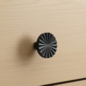 May include: A black, decorative cabinet knob with a radial design. The knob is mounted on a light-colored wooden surface. The knob's design features a series of radiating lines, creating a textured, sunburst-like appearance. The hardware is a stylish accent for furniture.