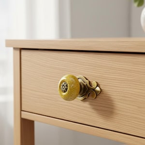 May include: A close-up of a wooden drawer with a decorative yellow and brass knob. The knob has a floral design in the center and is attached to the drawer with an ornate brass plate. The drawer is made of light-colored wood.