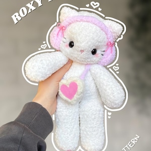 May include: A white plush cat toy with pink accents, wearing a pink headband and carrying a pink heart-shaped purse. The text "ROXY THE KITTY" is arched above the toy, and "DIGITAL PDF PATTERN" is below.