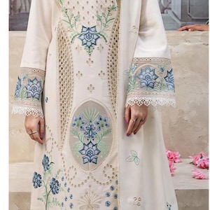 Può includere: Abito ricamato bianco sporco con maniche lunghe e sciarpa abbinata. L'abito presenta intricati motivi floreali blu e verdi, con dettagli in pizzo sulle maniche e sull'orlo. L'outfit è in stile tradizionale.