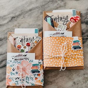 May include: Two brown paper packages, one wrapped in floral paper and the other in orange floral paper, tied with twine. Each package contains cards and stickers with the word "Fantasy" and the phrase "Blind Date With A Book".