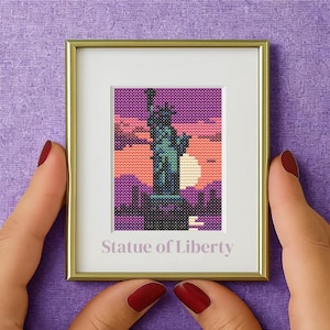 Peut inclure: Une œuvre d'art encadrée au point de croix représentant la Statue de la Liberté sur fond de coucher de soleil. L'œuvre est dans un cadre doré avec un passe-partout blanc. Le texte "Statue de la Liberté" est affiché en dessous.