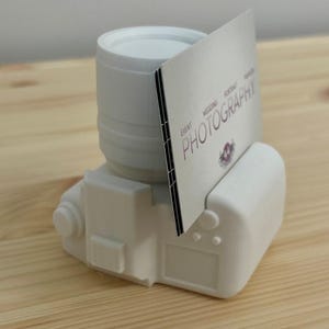 Könnte beinhalten: Ein weißer, 3D-gedruckter visitenkartenhalter in Kameraform. Der Halter hat ein Objektiv, einen Körper und einen Schlitz zur Aufnahme von Karten. Eine Visitenkarte mit dem Wort "FOTOGRAFIE" ist in den Schlitz eingesetzt. Die Karte enthält auch die Wörter "EVENT", "WEDDING", "PORTRAIT" und "FASHION".