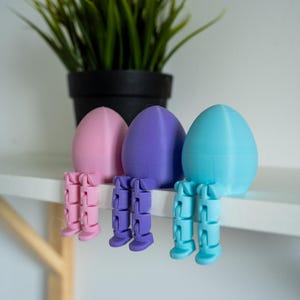 Può includere: Tre figurine colorate a forma di uovo con gambe articolate sedute su una mensola bianca. Le uova sono rosa pastello, viola e blu. Un vaso nero con erba verde sullo sfondo.