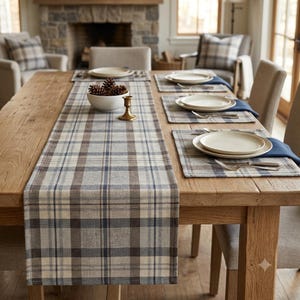 May include: A rectangular table runner featuring a plaid pattern in shades of beige, brown, and blue. The runner is laid across a wooden dining table, accompanied by matching placemats, white ceramic plates, silver cutlery, and a bowl of pinecones.