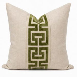 May include: A square throw pillow made of light beige linen fabric. The center features a vertical band of olive green velvet with a repeating geometric Greek key pattern, bordered by thin piping on both sides of the velvet strip.