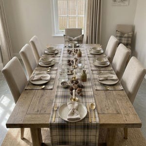 May include: A long rectangular wooden dining table set for eight with beige upholstered chairs. The table features a plaid runner, matching placemats, ceramic plates, gold-toned flatware, linen napkins, glass tumblers, and small candles.