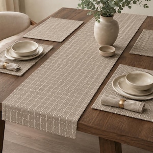 May include: A beige table runner and matching rectangular placemats featuring a white grid pattern. The set includes light-colored ceramic bowls and plates, linen napkins with gold-toned napkin rings, and a ceramic vase with greenery.