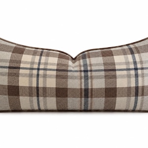 May include: A rectangular lumbar cushion featuring a brown, cream, and grey plaid pattern. The cushion has a distinct curved top edge and is finished with a dark brown piped trim along the entire perimeter, creating a structured and defined shape.