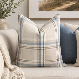 Peut inclure: Un coussin carr&eacute; avec un motif &agrave; carreaux tiss&eacute; dans des tons bleu clair, beige et blanc. Le tissu pr&eacute;sente un tissage textur&eacute; visible, et le motif se compose de lignes horizontales et verticales qui s'entrecroisent pour former une grille.