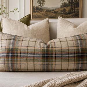 Peut inclure: Un coussin lombaire rectangulaire avec un motif à carreaux dans des tons de beige, vert olive et bleu marine. Derrière, deux coussins carrés en tissu beige texturé et deux coussins carrés en velours vert olive uni sont disposés.