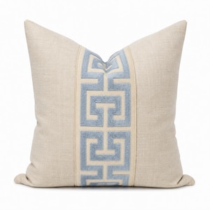 May include: A square throw pillow in a light beige, textured fabric. The center features a vertical band of light blue velvet embroidery in a geometric Greek key pattern, framed by thin, raised light beige fabric borders on both sides.