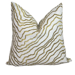 May include: A decorative throw pillow with a white background and a gold, wavy pattern. The pillow has a textured appearance and a soft, inviting look. The pillow is designed for home decor.