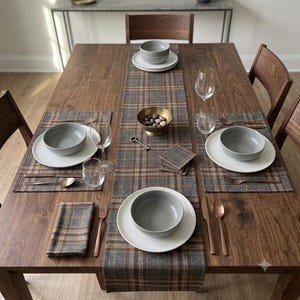 May include: A dark wood dining table set for three with a plaid table runner and matching placemats. Each place setting includes a white plate, a grey bowl, a clear glass, and copper-toned cutlery. A brass bowl with stones sits in the centre.