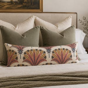 May include: A rectangular lumbar pillow featuring a repeating fan-shaped botanical pattern in shades of muted pink, mustard yellow, and dark charcoal on a cream background, positioned in front of two olive green square pillows and two cream pillows.