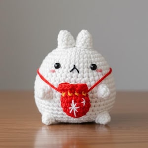 Op de afbeelding: Een handgemaakt wit gehaakt konijn met zwarte ogen, een kleine zwarte neus en roze wangen. Het heeft een rode buidel met een wit sneeuwvlokontwerp. Het konijn zit op een houten oppervlak.