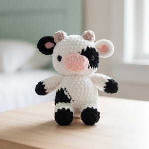 May include: A handmade crochet cow plushie. The stuffed animal is white with black spots, black hooves, and black ears with pink inner ears. It has a pink nose and small beige horns. The cow is sitting on a light brown wooden table.