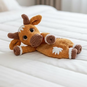 Può includere: Un peluche a forma di mucca all'uncinetto fatto a mano, nei toni del marrone e dell'arancione, con accenti bianchi. La mucca è sdraiata su un copriletto bianco. Il giocattolo ha grandi occhi espressivi ed è realizzato con filato morbido e strutturato.