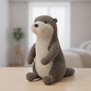 May include: A crocheted otter toy in shades of grey and cream. The otter has a detailed design with a cream-colored face and belly, and dark grey body. It is sitting on a light wooden surface, with a blurred background.