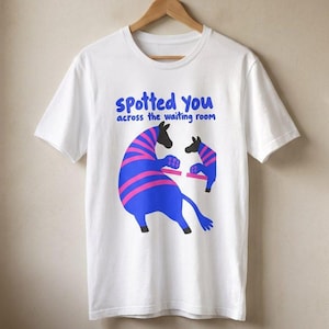 May include: A white short-sleeved t-shirt featuring a graphic of two blue and pink striped zebras facing each other. Above the zebras, the text reads "spotted you across the waiting room" in blue letters. The shirt is displayed on a wooden hanger.