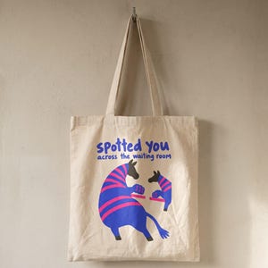 May include: A beige canvas tote bag featuring the blue text "spotted you across the waiting room" above a graphic of two blue and pink striped animals facing each other. The bag has two long shoulder straps and a rectangular body shape.