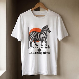May include: A white short-sleeve t-shirt featuring a graphic of a zebra standing in front of an orange circle. Below the zebra are five crescent moon icons and the text "hoofbeats. finally, zebras." The shirt is displayed on a wooden hanger.