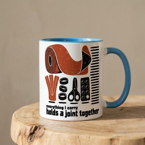 May include: A white ceramic mug with a blue interior and handle, featuring a graphic print of a roll of tape, scissors, and other items. The text printed on the mug reads: everything i carry holds a joint together in bold black lettering.
