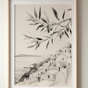 以下が含まれることがあります： 海岸沿いの村の風景を描いた、額入りの白黒プリント。 オリーブの実が付いたオリーブの枝が前景にあります。 村にはドーム型の屋根を持つ白い建物があります。 アートワークは明るい木製のフレームに入っています。