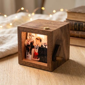 Peut inclure: Une boîte de présentation photo carrée en bois avec une finition à grain brun foncé. L'ouverture avant révèle une photographie de deux personnes s'enlaçant. Une petite manivelle en bois est fixée sur le côté droit et un bouton en laiton est sur le dessus.