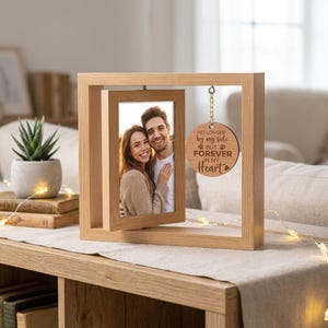 Peut inclure: Un cadre photo rotatif en bois clair contenant une photographie d'un couple. Un ornement circulaire en bois est suspendu au cadre supérieur, gravé du texte : NO LONGER by my side BUT FOREVER IN MY Heart, accompagné de deux empreintes de pattes.